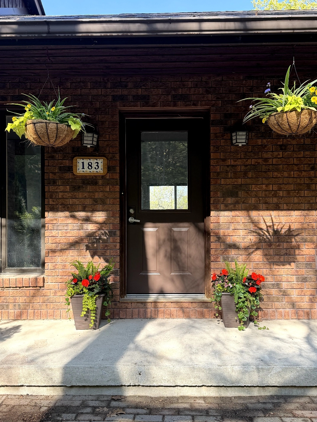 Front door with brick facade, hanging flower baskets, and potted plants